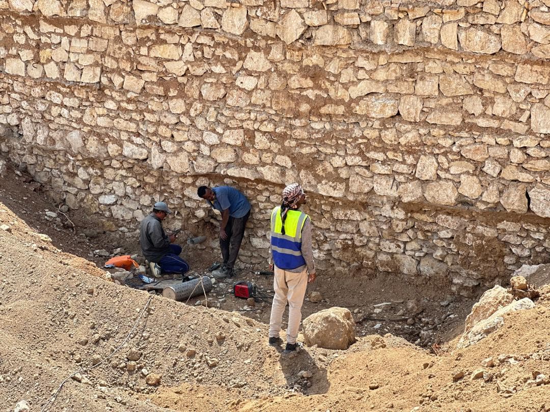 Workers inspecting pipework beside a stone infrastructure wall